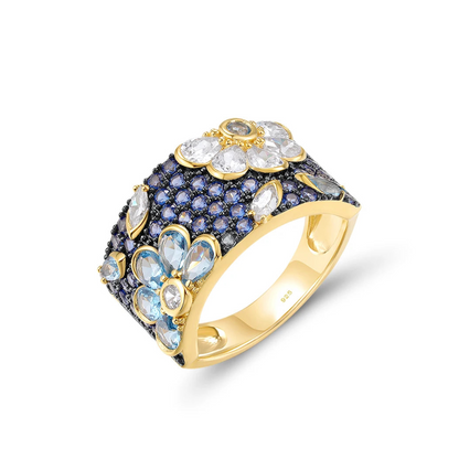 Gold-plated floral ring with a wide band, set with clear, blue, and navy gemstones forming flower shapes

