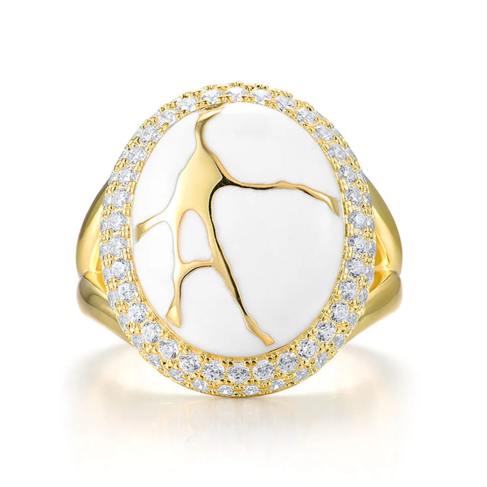 Gold ring with a large white stone featuring golden vein-like patterns, encircled by two rows of clear stones.

