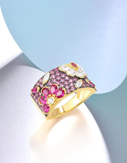 Wide gold ring adorned with vibrant pink and white gemstones arranged in floral patterns.

