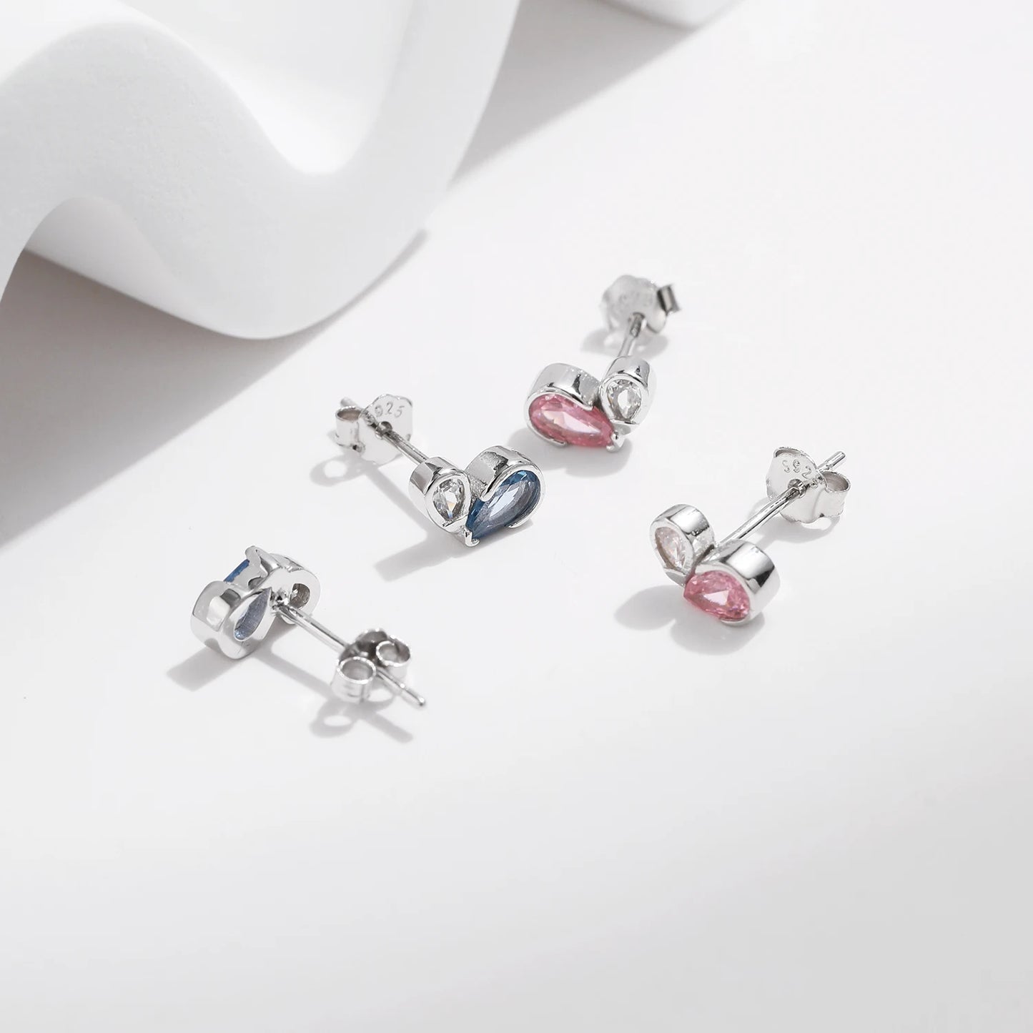 Pair of silver earrings featuring a pink or blue heart-shaped gemstone paired with a smaller round clear stone in a modern open setting.

