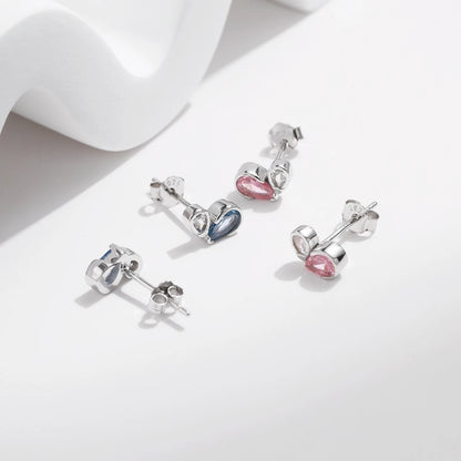 Pair of silver earrings featuring a pink or blue heart-shaped gemstone paired with a smaller round clear stone in a modern open setting.

