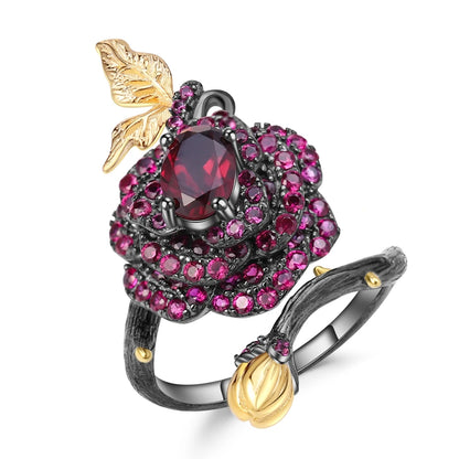 Black rose ring with layered red gemstones, accented by a gold leaf and bud on a dark branch-like band.

