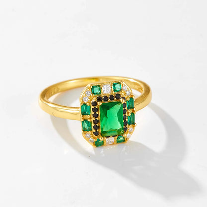 Gold ring with a central emerald-green rectangular stone, surrounded by alternating black and white crystals and square-cut green gems.

