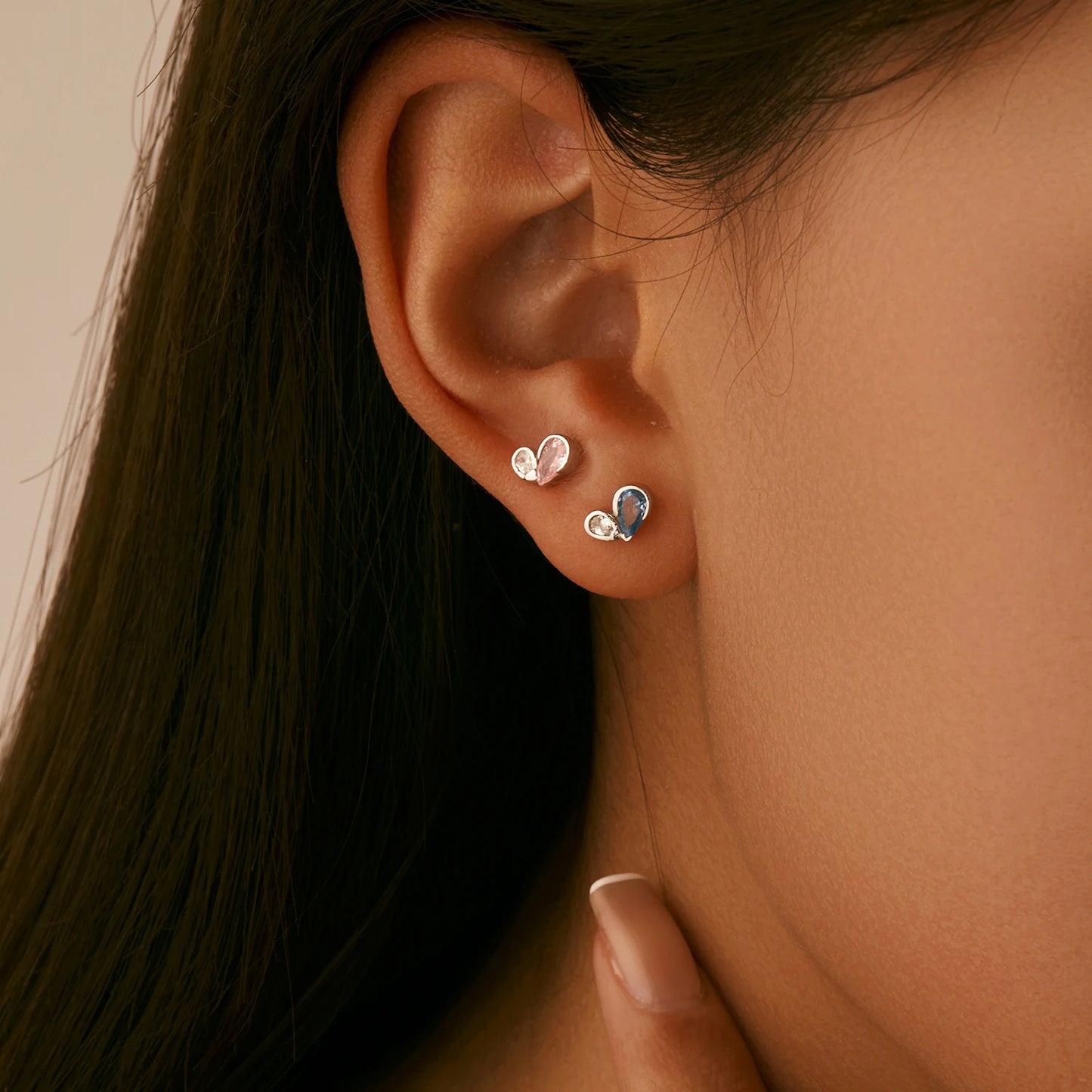 Pair of silver earrings featuring a pink or blue heart-shaped gemstone paired with a smaller round clear stone in a modern open setting.

