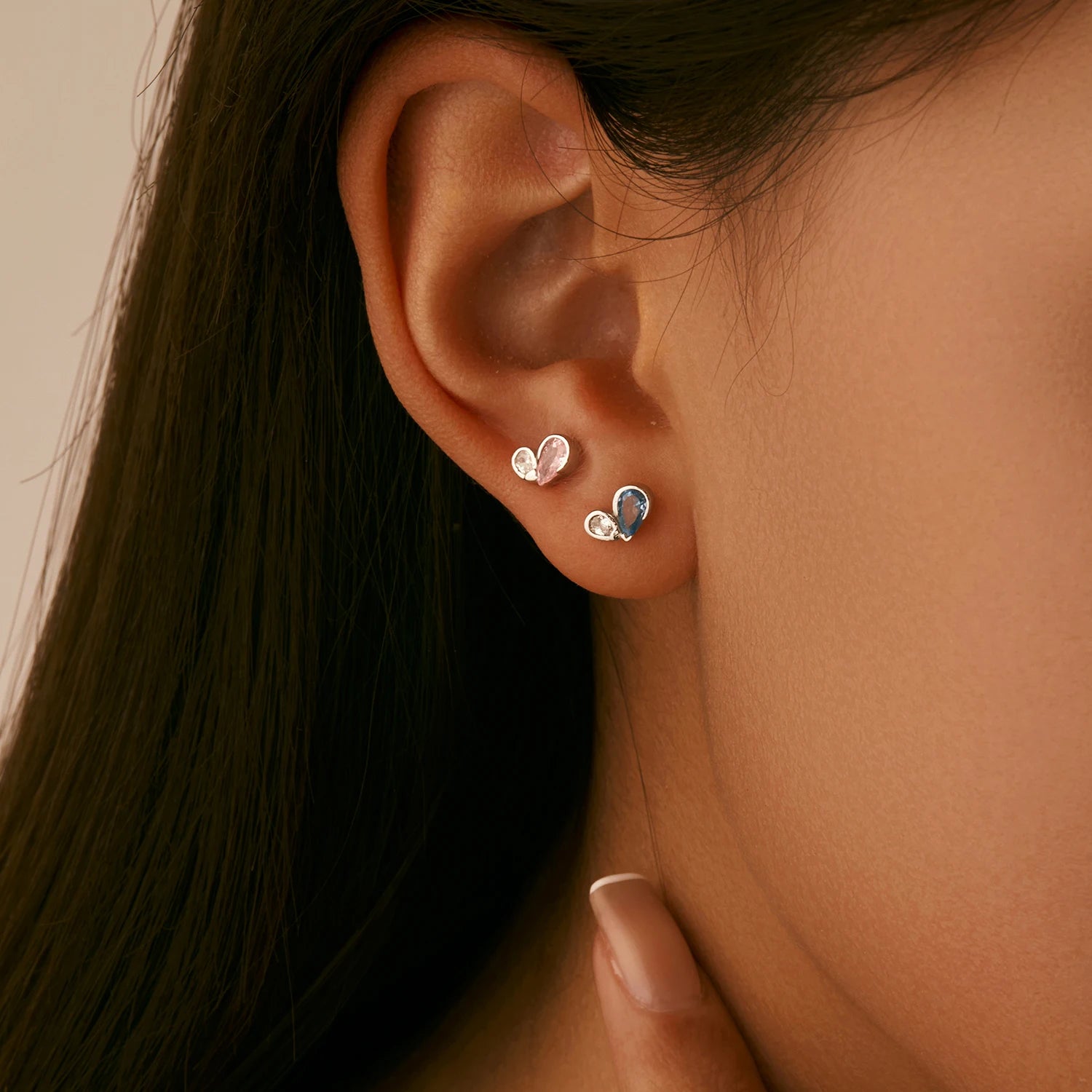 Pair of silver earrings featuring a pink or blue heart-shaped gemstone paired with a smaller round clear stone in a modern open setting.

