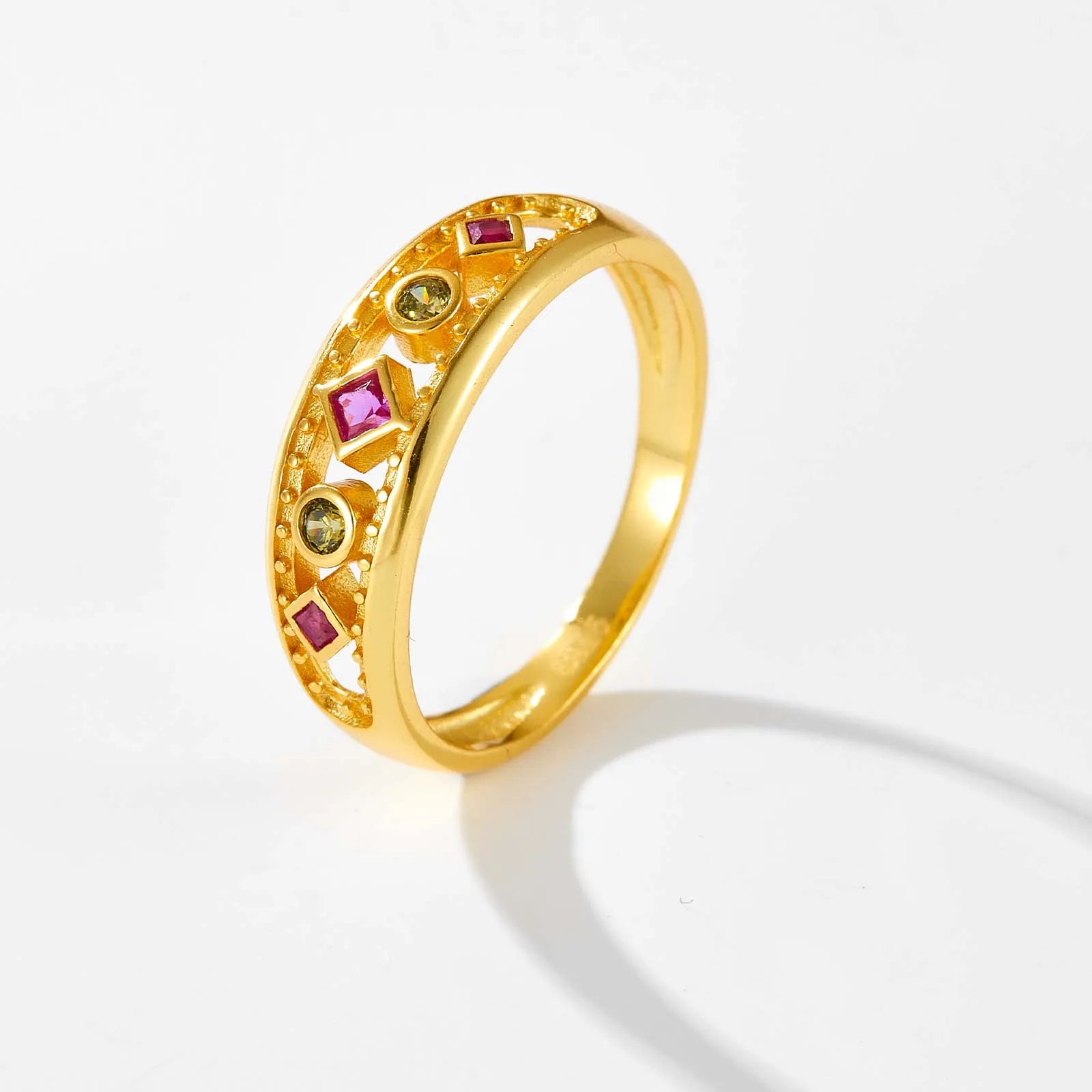 Gold ring with alternating pink square-cut and green round gemstones, framed by beaded detailing, displayed on a red background.

