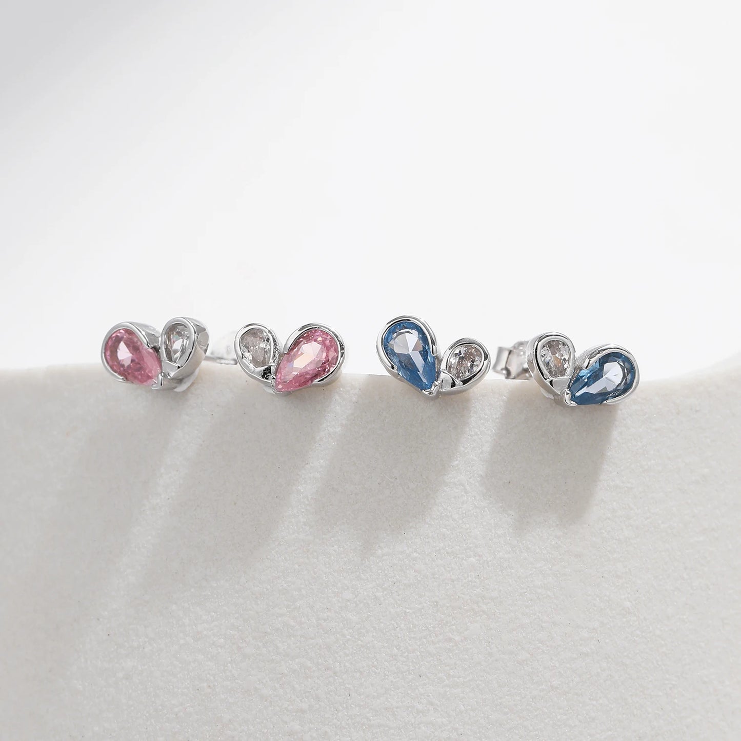 
Pair of silver earrings featuring a pink or blue heart-shaped gemstone paired with a smaller round clear stone in a modern open setting.



