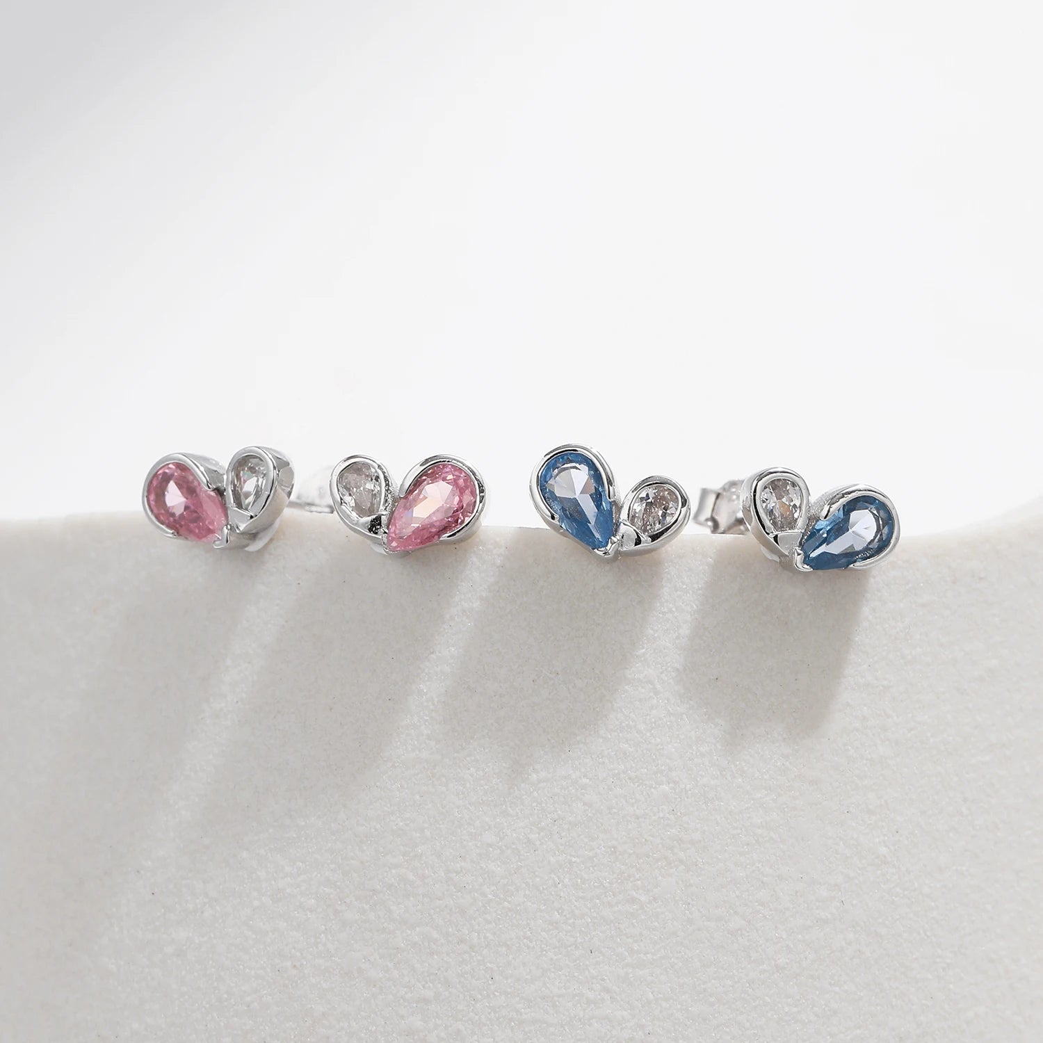 
Pair of silver earrings featuring a pink or blue heart-shaped gemstone paired with a smaller round clear stone in a modern open setting.



