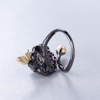 Black rose ring with layered red gemstones, accented by a gold leaf and bud on a dark branch-like band.

