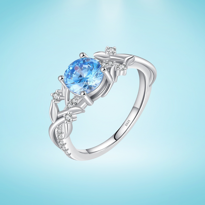 Sterling silver ring with a round sky blue gemstone and flowing leaf-like band details accented by small clear stones, set on a light blue background