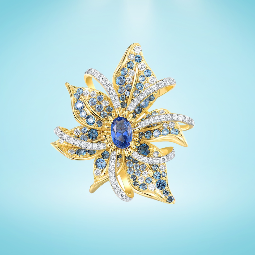 Gold floral brooch with layered petal-like curves, set with blue gemstones and clear pavé accents, featuring an oval sapphire-blue centerpiece