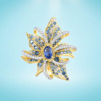 Gold floral brooch with layered petal-like curves, set with blue gemstones and clear pavé accents, featuring an oval sapphire-blue centerpiece