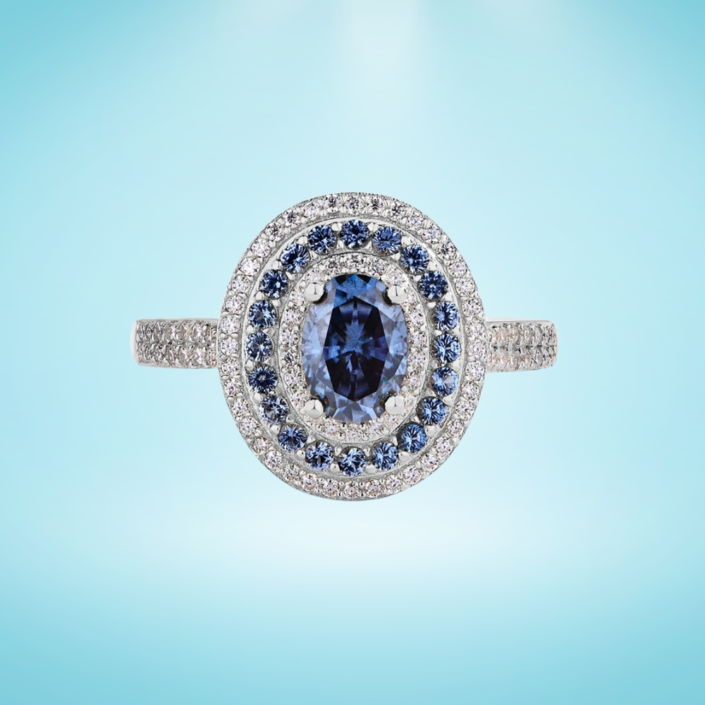 Sterling silver ring with an oval-cut deep blue gemstone surrounded by two concentric halos of white and blue stones, set on a pavé band, on a light blue background