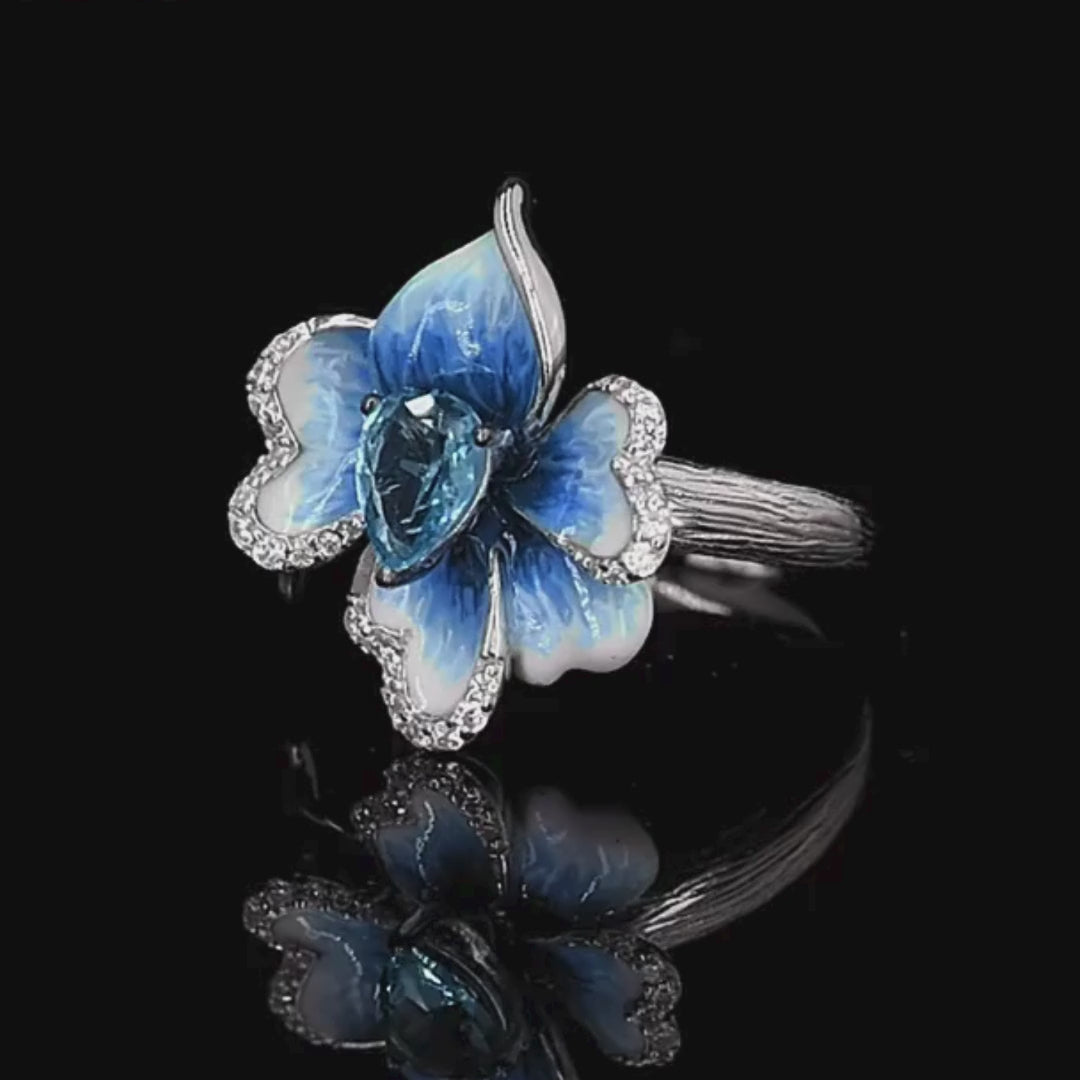 Silver ring with a sculpted blue enamel flower and a central teardrop-shaped blue gemstone surrounded by white stones

