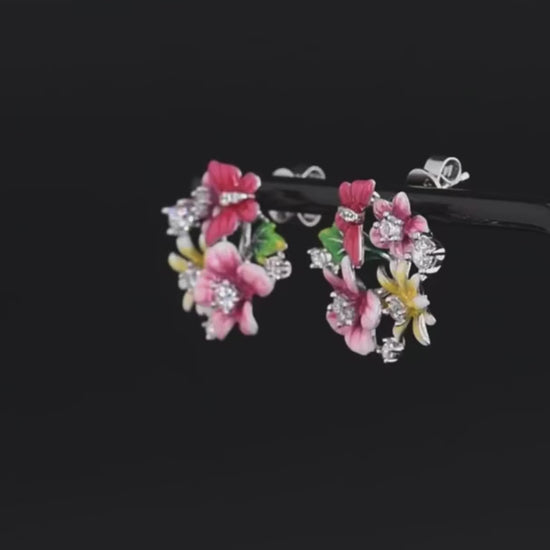 Pair of vibrant floral stud earrings with pink, red, and yellow enamel flowers accented by sparkling stones, set against a red background.

