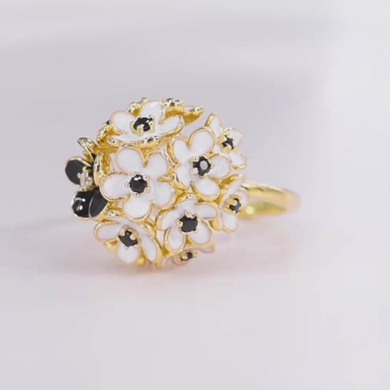 Gold ring featuring a cluster of white and black enamel flowers with dark gemstone centers.

