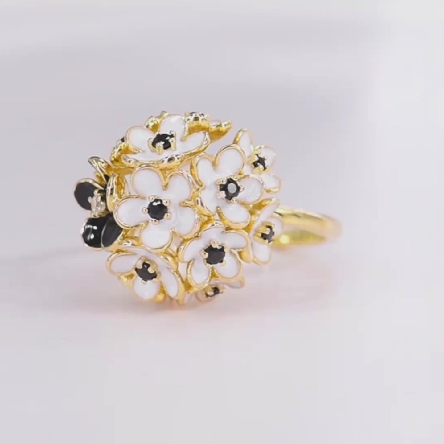Gold ring featuring a cluster of white and black enamel flowers with dark gemstone centers.

