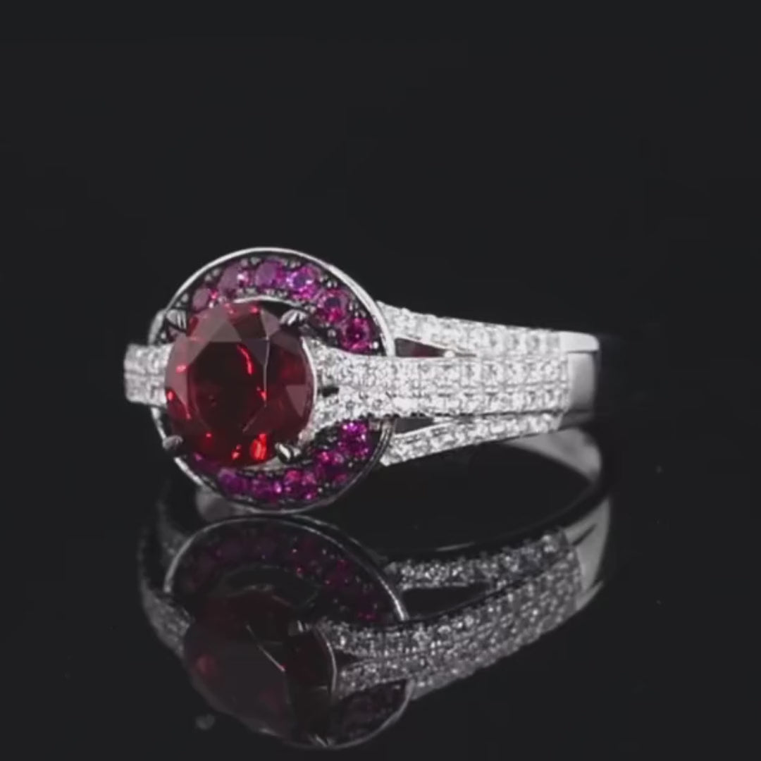 Silver ring with a large round red center stone, surrounded by a crescent of smaller pink stones and pavé white accents on a split band.

