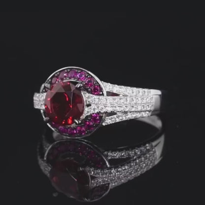 Silver ring with a large round red center stone, surrounded by a crescent of smaller pink stones and pavé white accents on a split band.

