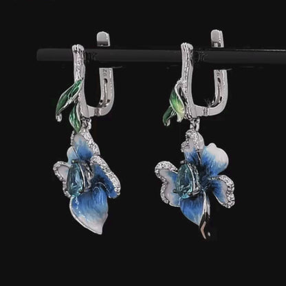 Silver floral earrings with hand-painted blue petals, green enamel accents, and teardrop blue stones.

