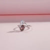 Silver ring with a split band featuring a clear pear-cut stone and a deep rose oval gem, shown on a red background from the Rossa collection.

