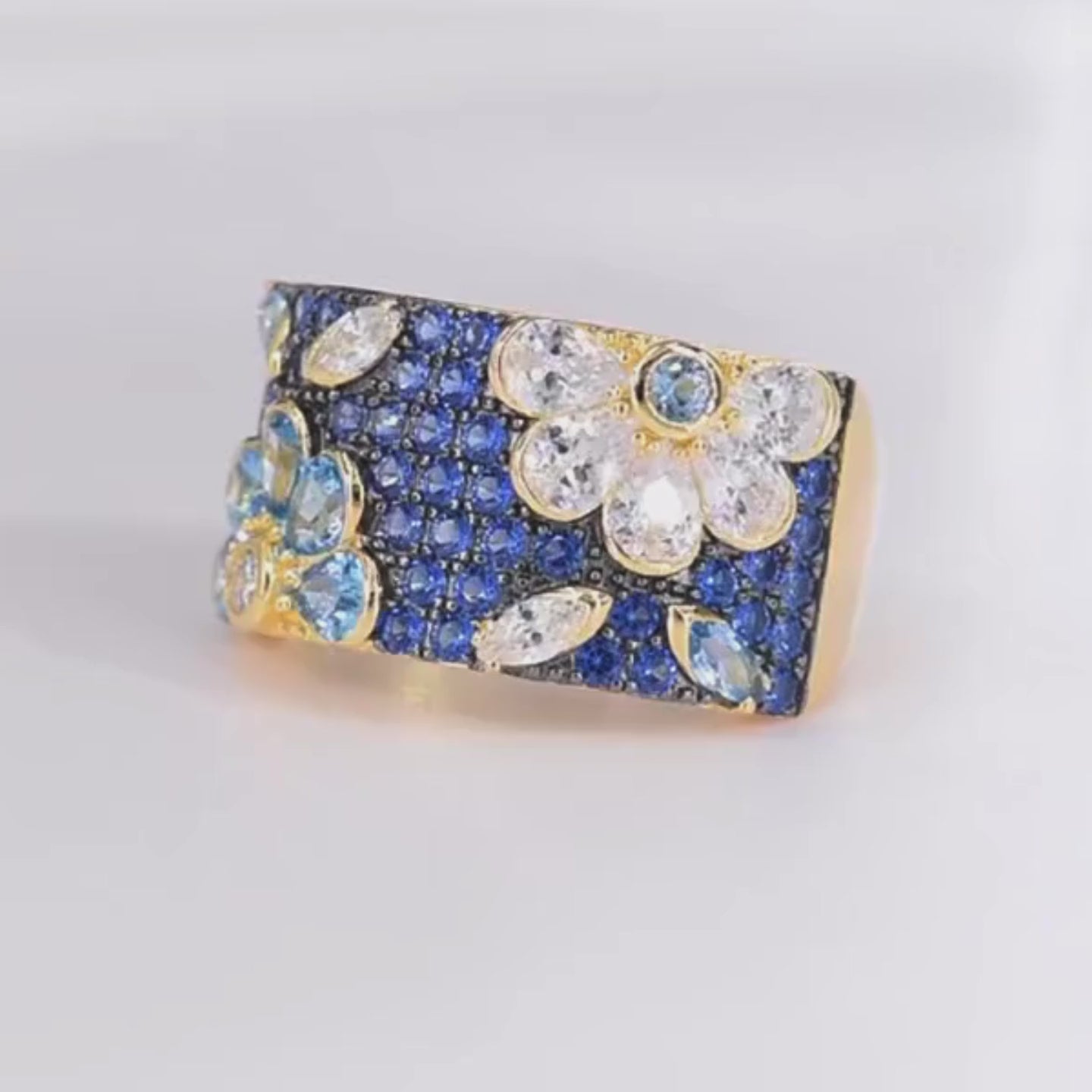 Gold-plated floral ring with a wide band, set with clear, blue, and navy gemstones forming flower shapes

